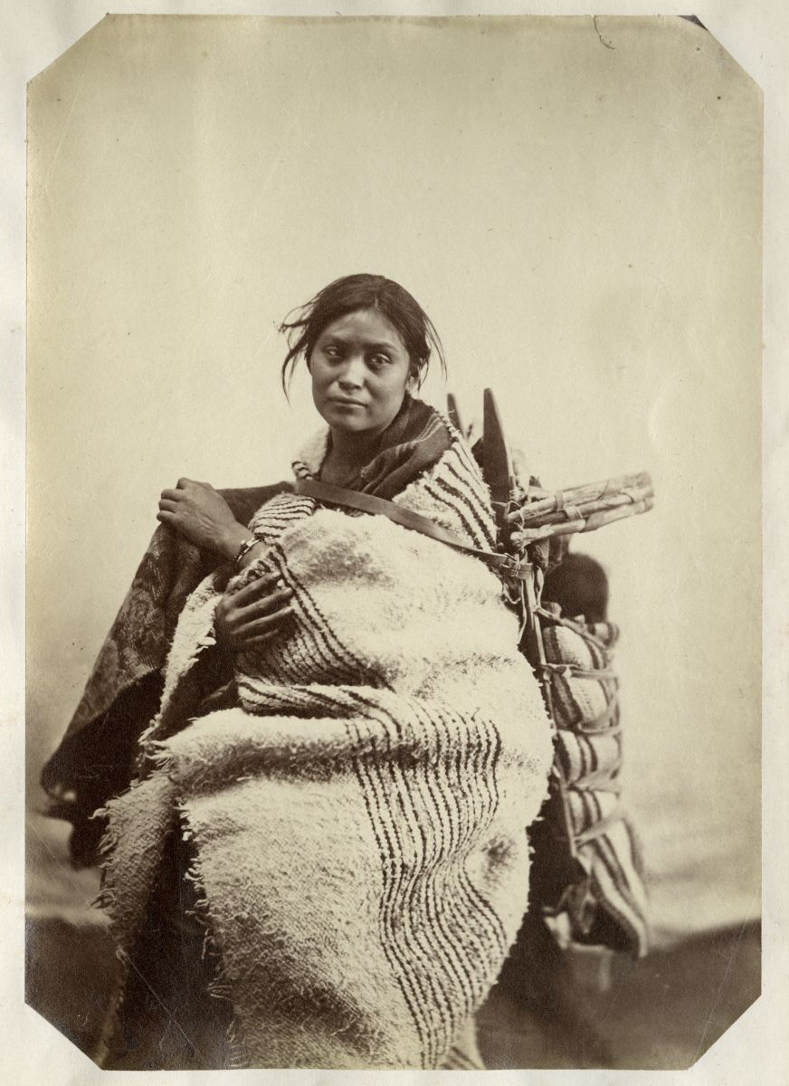 A sepia-toned photograph shows a Dine, or Navajo, woman wearing a blanket and carrying an infant on her back in a cradleboard